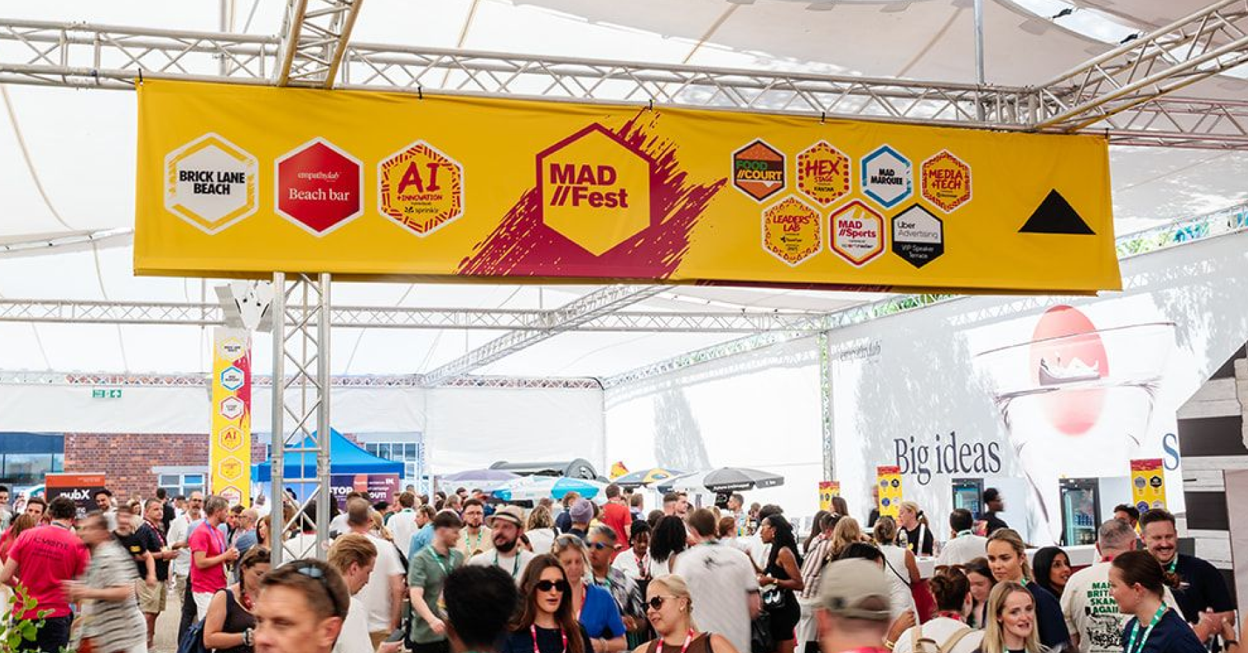 A busy outdoor crowd of marketing professionals at the MAD//Fest London conference venue underneath a large yellow directional sign featuring event stages.